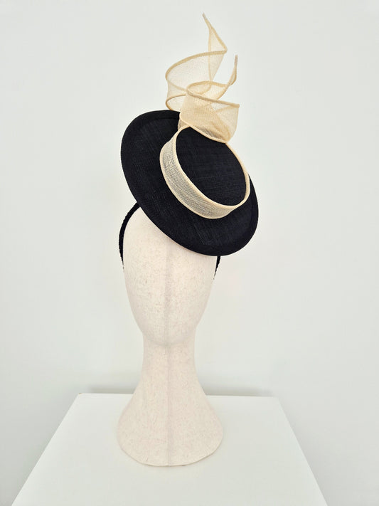 A black straw percher hat fascinator with cream swirl details and  band  displayed on a textured cream-colored mannequin head against a white studio background.