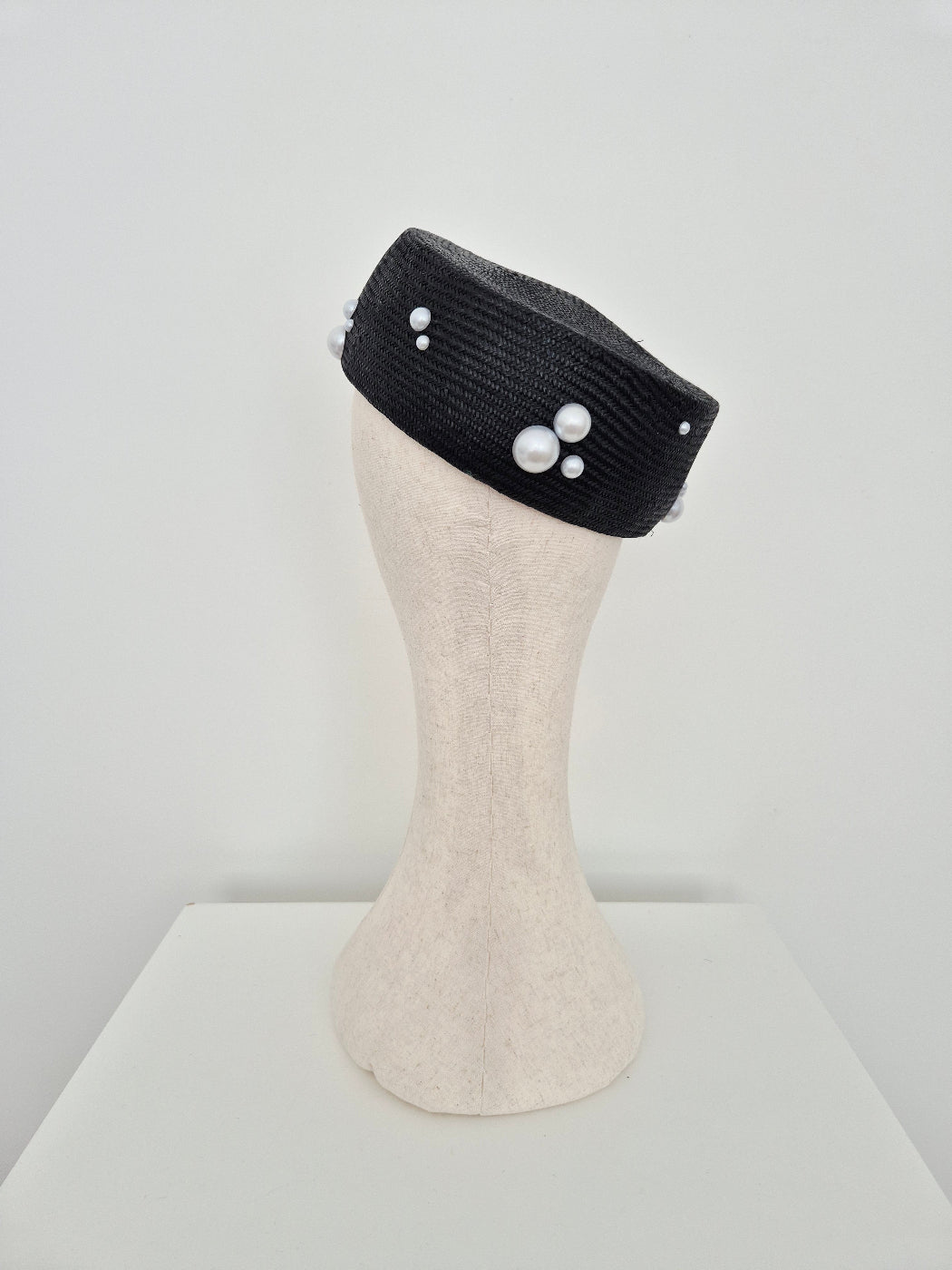 A black straw pillbox style hat with white faux-pearl embellishments, displayed on a cream-colored mannequin head against a light gray background.back view
