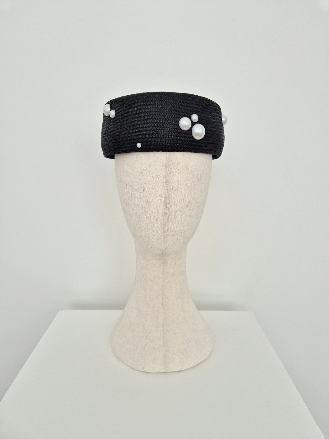 A black straw headpiece with white faux-pearl embellishments, displayed on a cream-colored mannequin head against a plain white background.