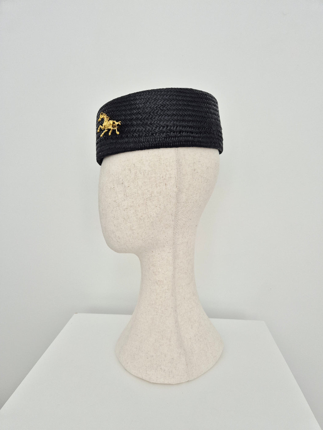 black straw pillbox headpiece with gold horse brooch side view displayed on a manniquin head
