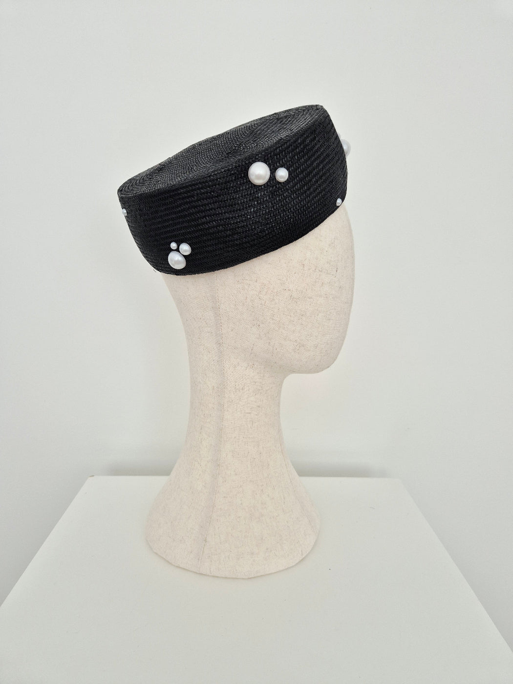 A black straw pillbox style headpiece with white decorative pearl-like accents, displayed on a cream-colored mannequin head against a plain white background.side view