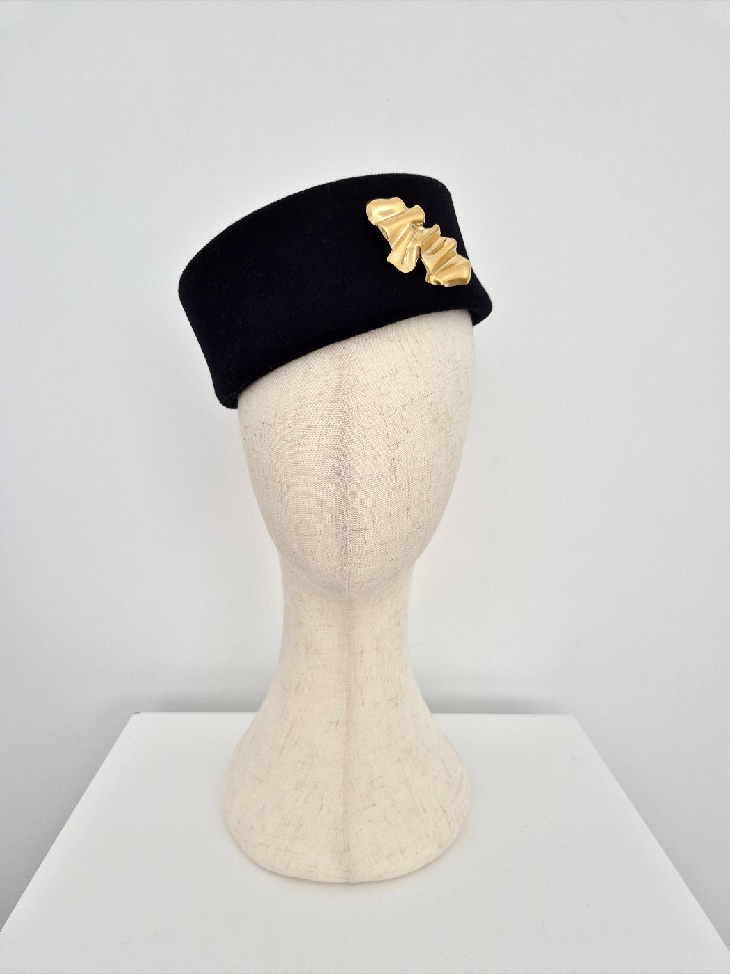 Black pillbox headpiece  with gold brooch on a mannequin head against a white background