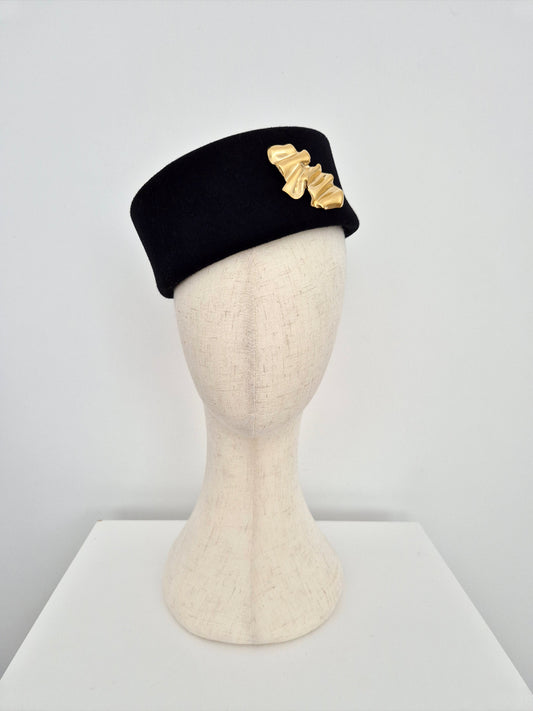 Black pillbox headpiece  with gold brooch on a mannequin head against a white background