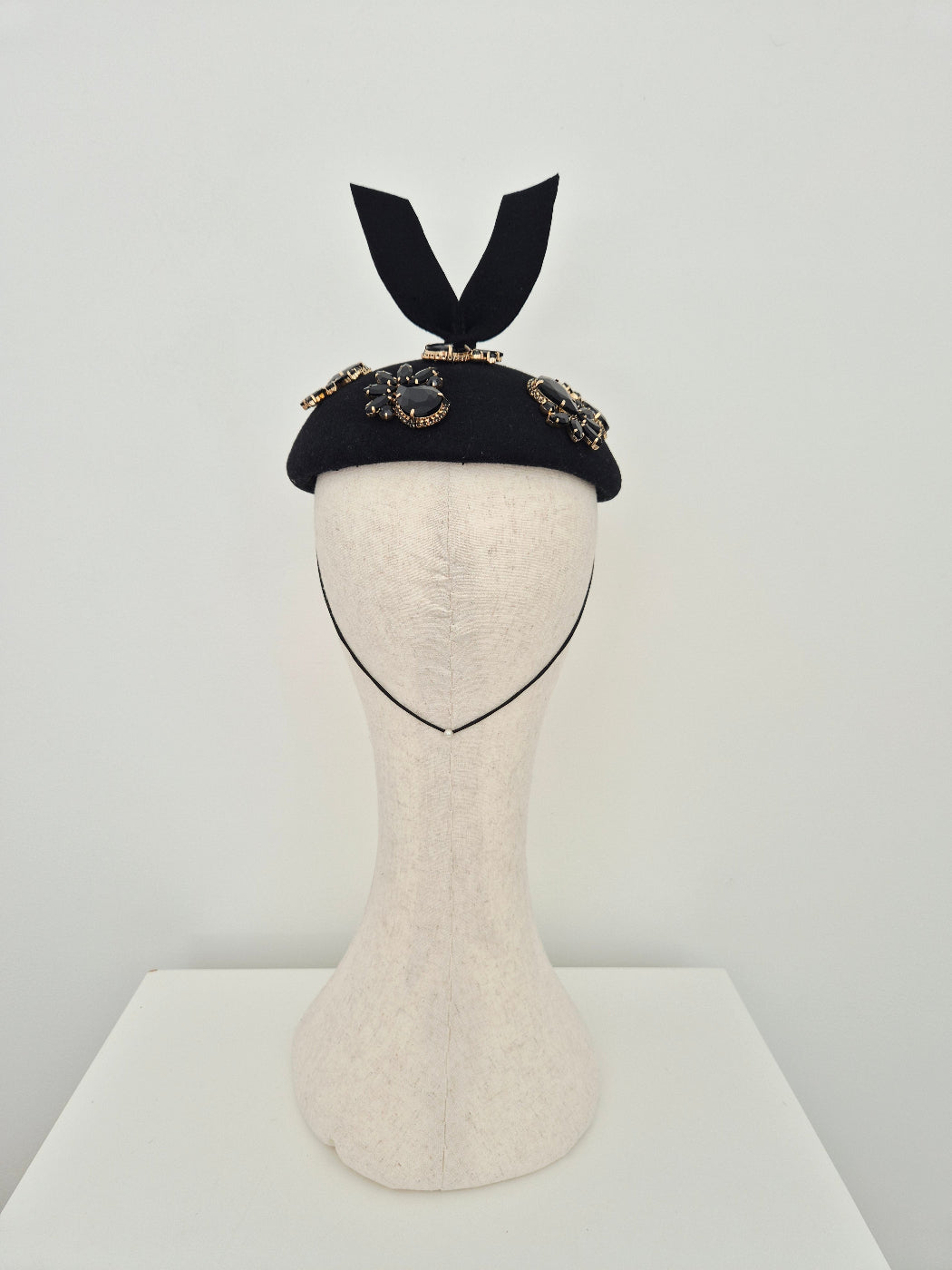 A black felt button fascinator hat with ornate rhinestone embellishments and a black ribbon bow, displayed on a cream mannequin head against a light gray background. back view