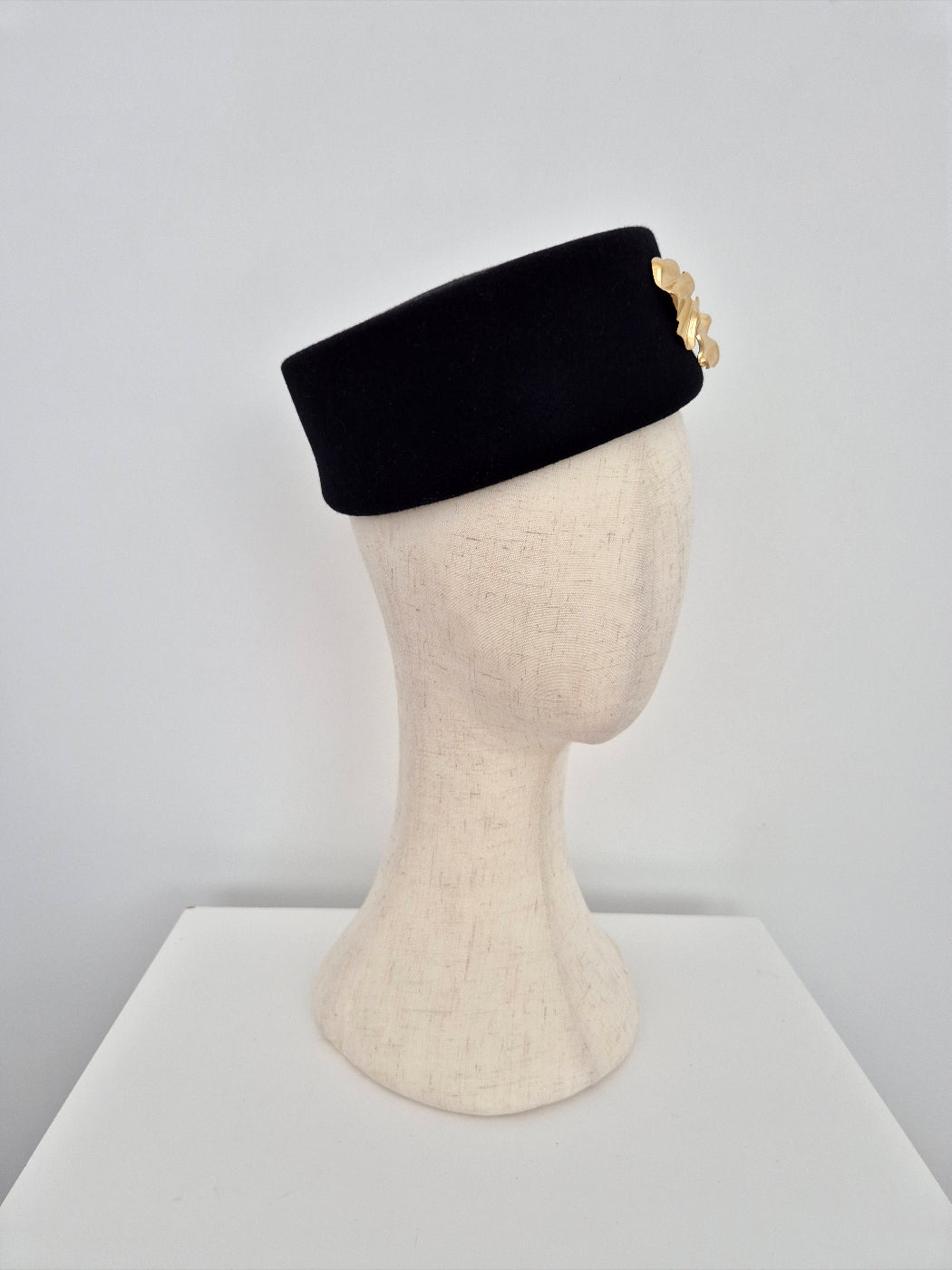 Black pillbox headpiece with gold brooch on a mannequin head against a gray background