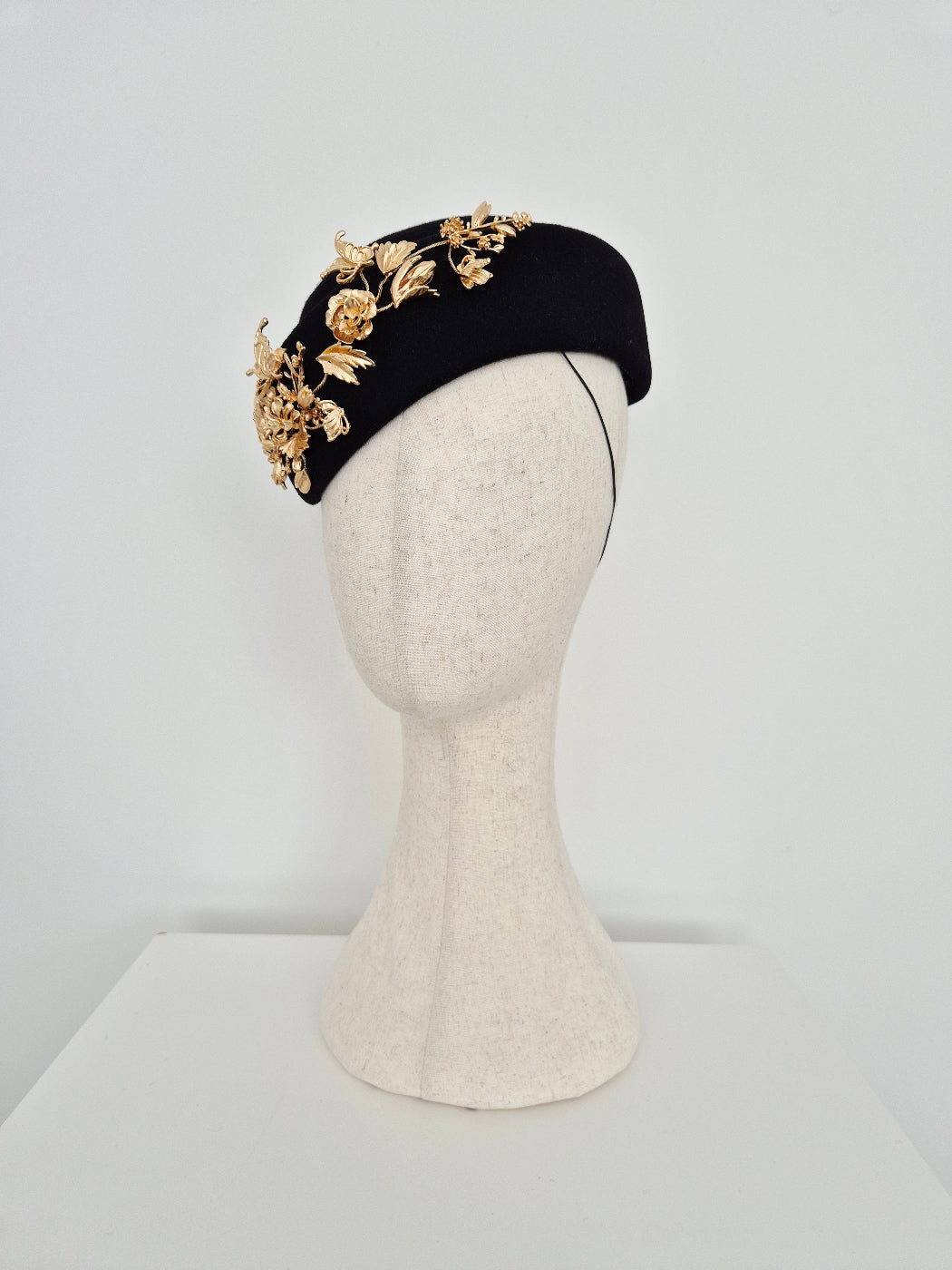 Black headpiece hostess style in wool felt with gold floral embellisments on a mannequin head against a white background side view