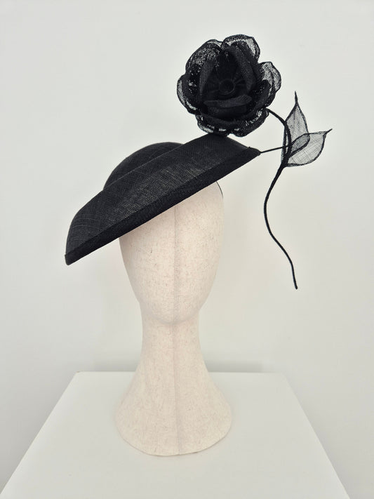 A black fascinator hat with a matching black mesh rose and leaf accent, displayed on a cream-colored mannequin head against a white background.