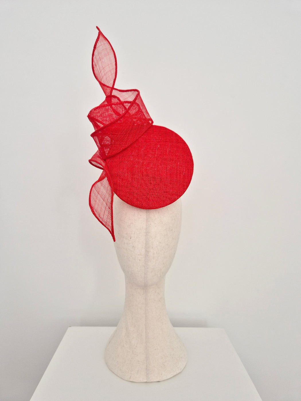 A bright red fascinator hat displayed on a textured off-white mannequin head against a plain light gray background.