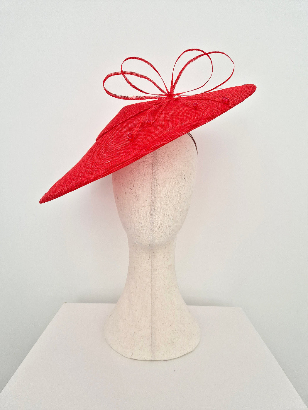 A bright red fascinator hat with a matching red ribbon bow, displayed on a textured off-white mannequin head against a plain white background.