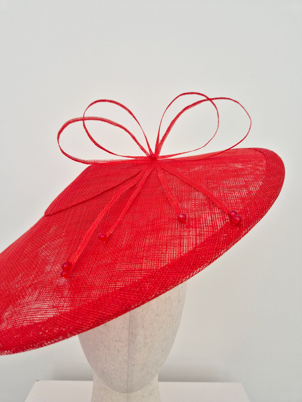 A bright red wide-brimmed fascinator hat with a matching red ribbon bow and small bead accents, displayed on a light gray mannequin head against a plain light background.close up