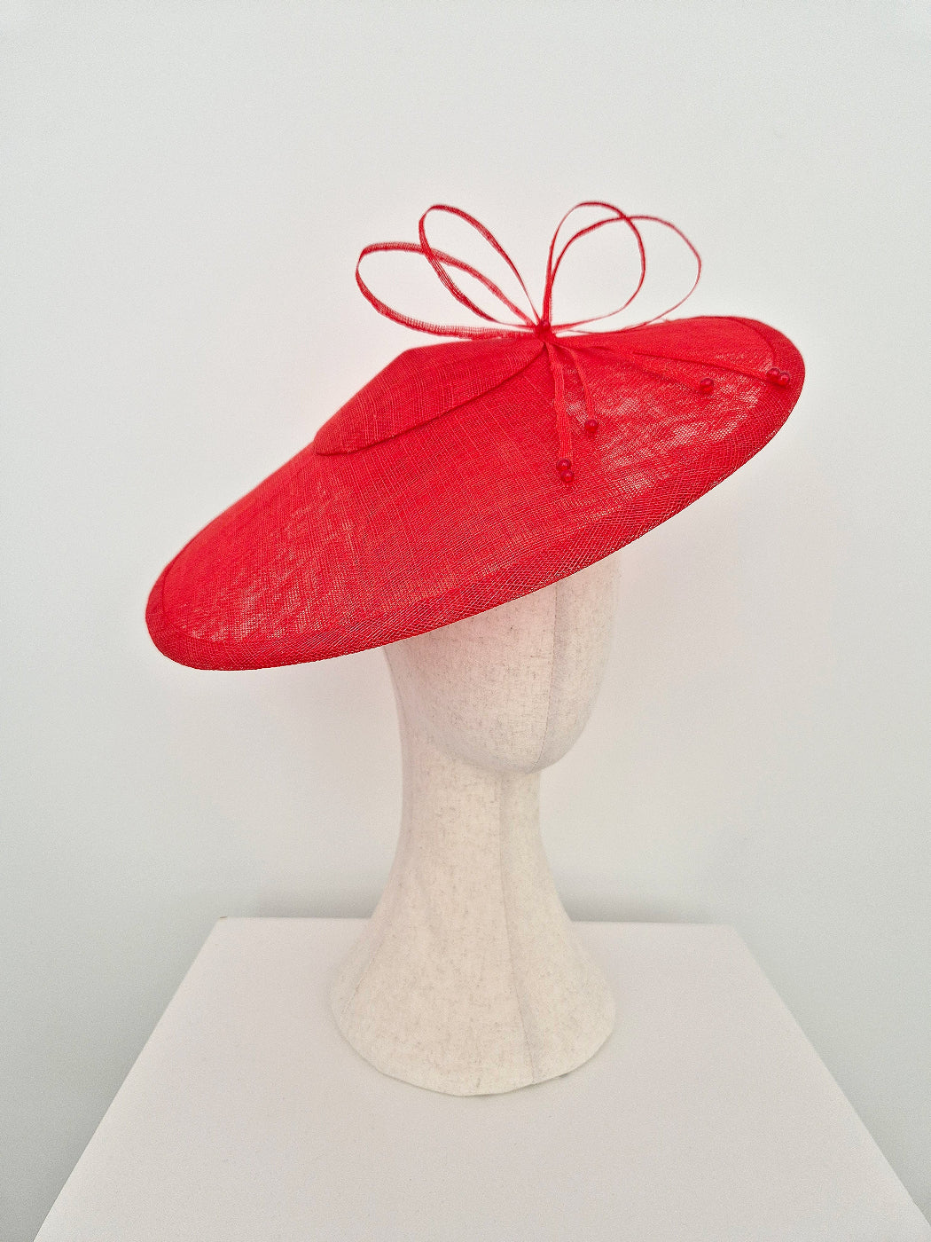 A bright red wide-brimmed fascinator hat with a matching red ribbon bow on top, displayed on a cream-colored mannequin head against a plain white background. side view