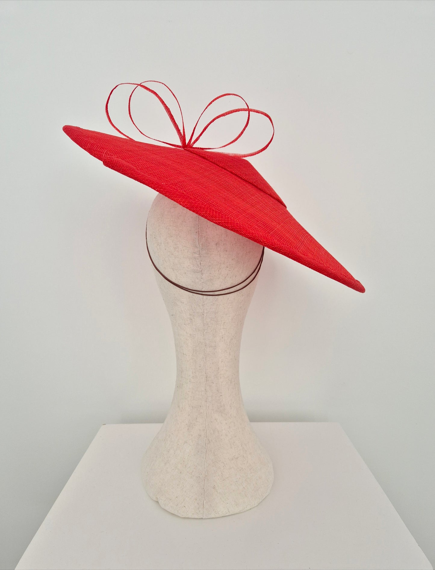 A bright red fascinator hat with a wide, brim and a matching red ribbon loop bow on top, displayed on a cream-colored mannequin head against a plain white background.back view