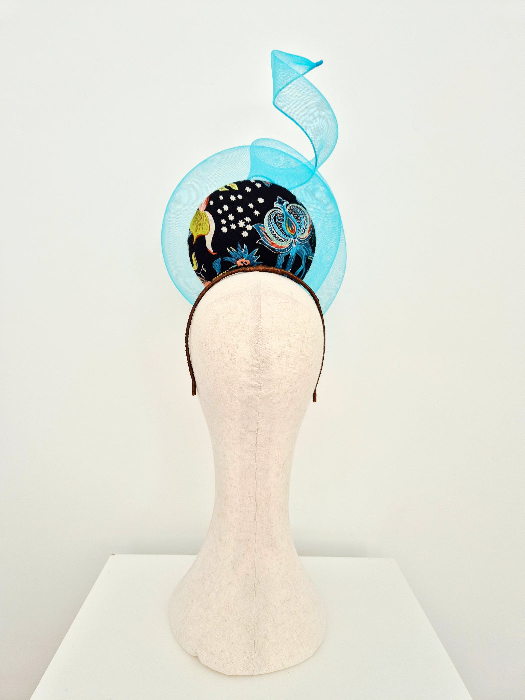 A decorative fascinator hat displayed on a cream fabric mannequin head against a plain white studio background. The hat features a blue crinoline swirl section, a black patterned lower section with colorful abstract motifs, back view