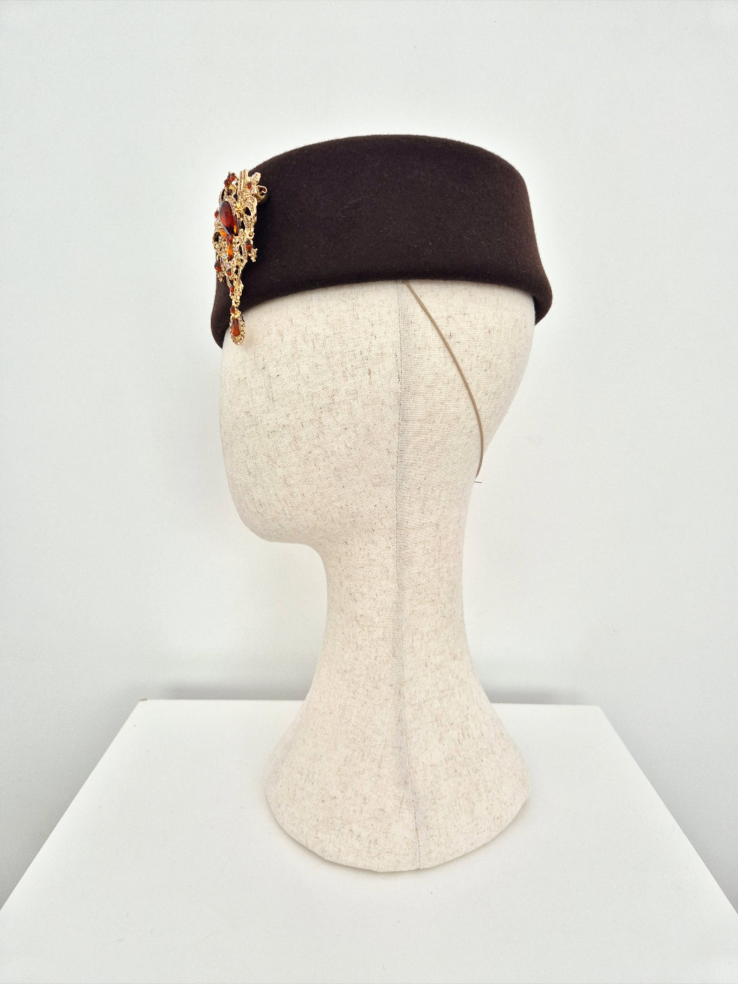 Brown pillbox headpiece with brooch on a mannequin head against a white background side view