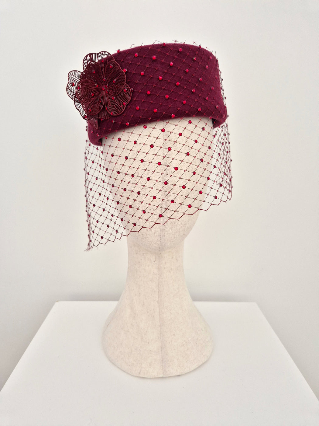 burgundy felt pillbox headpiece with a floral detail and netting on a mannequin head against a white background