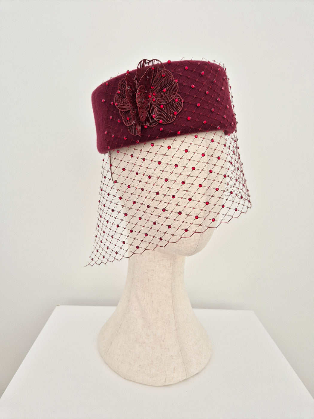 Burgundy pillbox hat with a net veil on a mannequin head against a white background side view