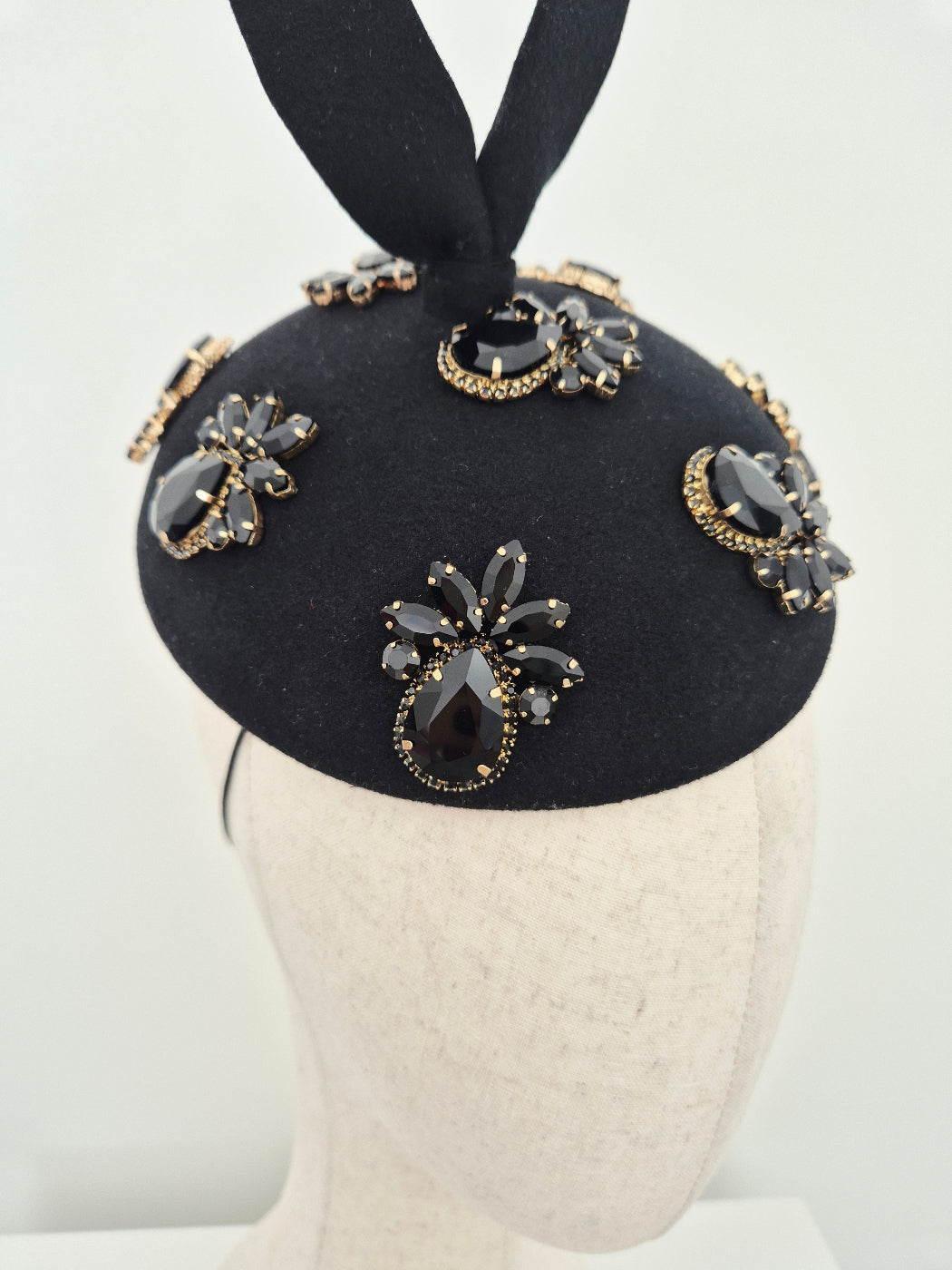 A close-up of a black felt fascinator headband with black and gold rhinestone brooches and a black ribbon tail, displayed on a cream-colored mannequin head against a white background.