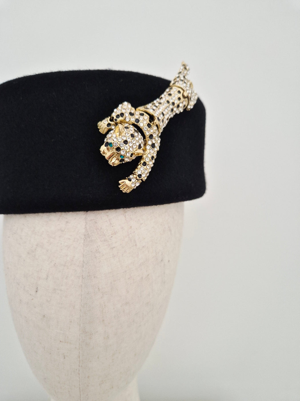 Gold leopard detail on a black felt pillbox headpiece on a mannequin head against a white background close up