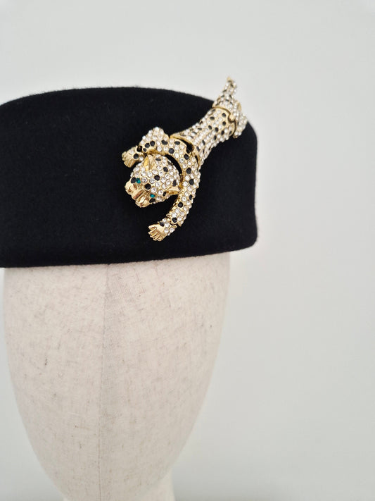 Gold leopard detail on a black felt pillbox headpiece on a mannequin head against a white background close up