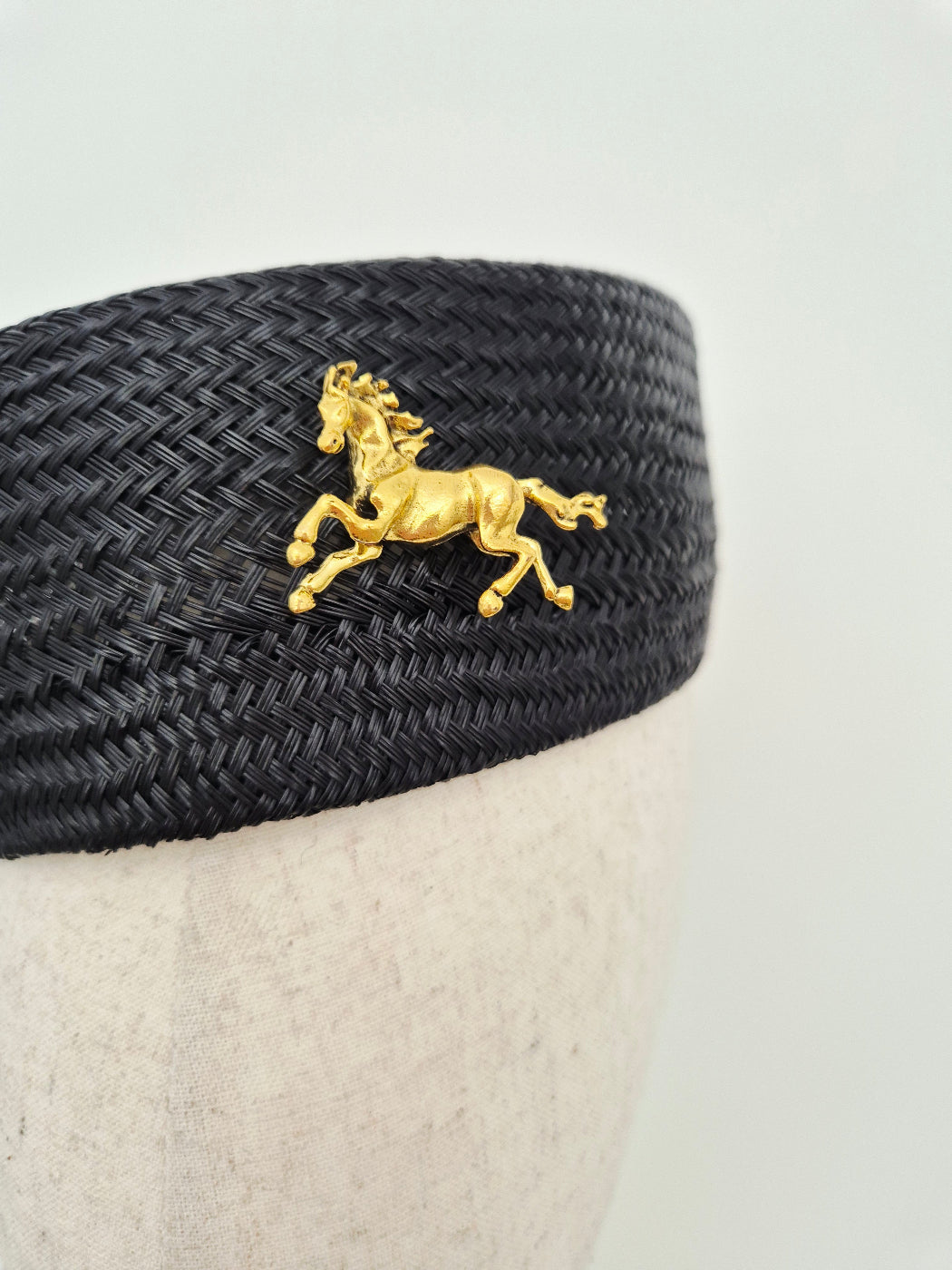 A gold horse-shaped brooch displayed on a black woven hat band, mounted on a light beige fabric mannequin head against a plain light gray background.close up