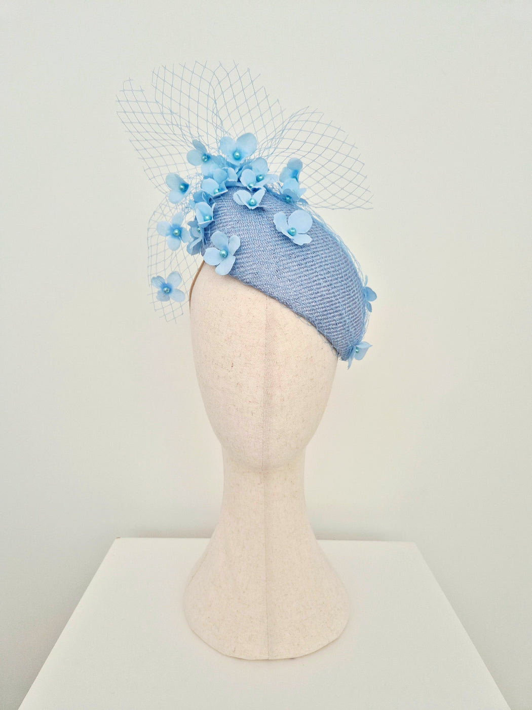 A light blue textured fascinator  with light blue fabric flowers and a white birdcage veil, displayed on a beige mannequin head.