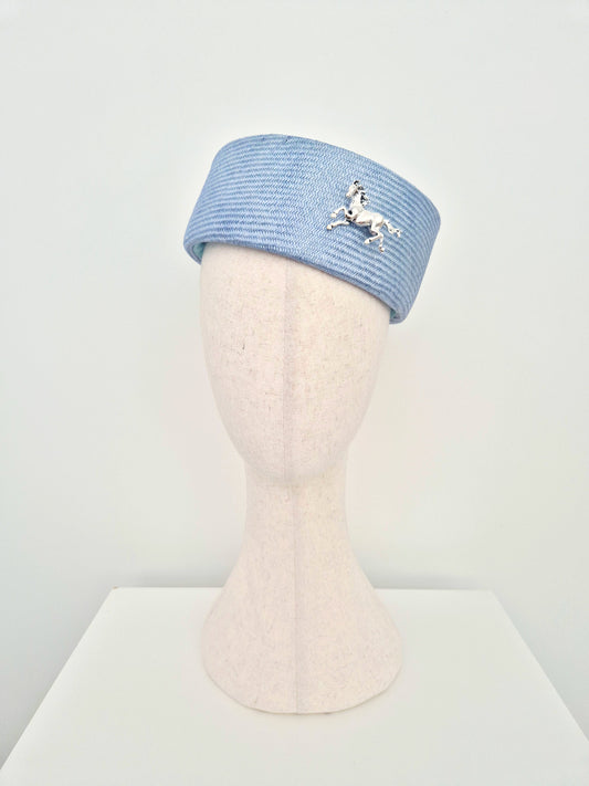A dusty blue straw pillbox hat with a silver horse brooch, displayed on a cream-colored mannequin head against a white background. tilted