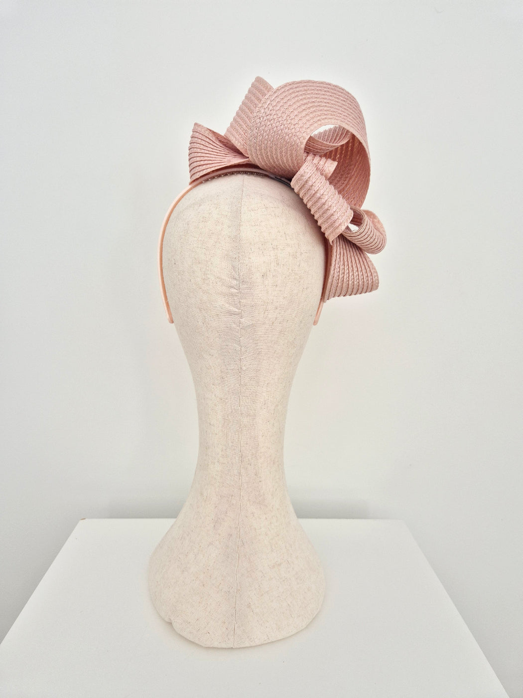 A dusty pink pleated fascinator hat with a large bow, displayed on a cream-colored felt mannequin head against a white background.back view