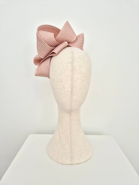 A dusty pink woven fascinator hat with a large bow, displayed on a cream-colored felt mannequin head against a white background.