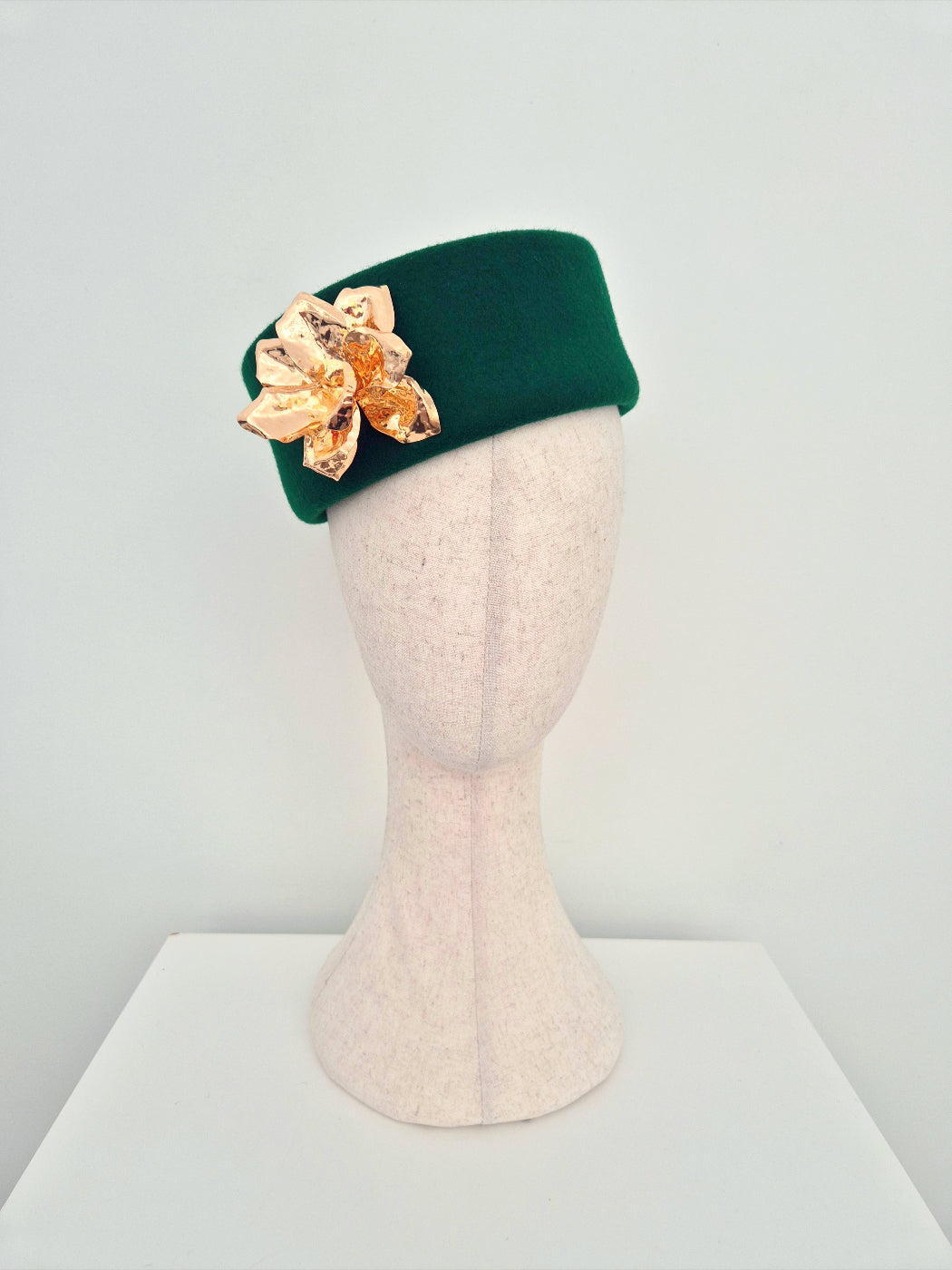 Green pillbox hat with a gold flower on a mannequin head against a white background