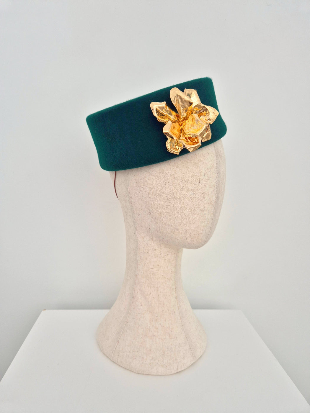 Green headpiece pillbox with a gold flower on a mannequin head against a white background side view