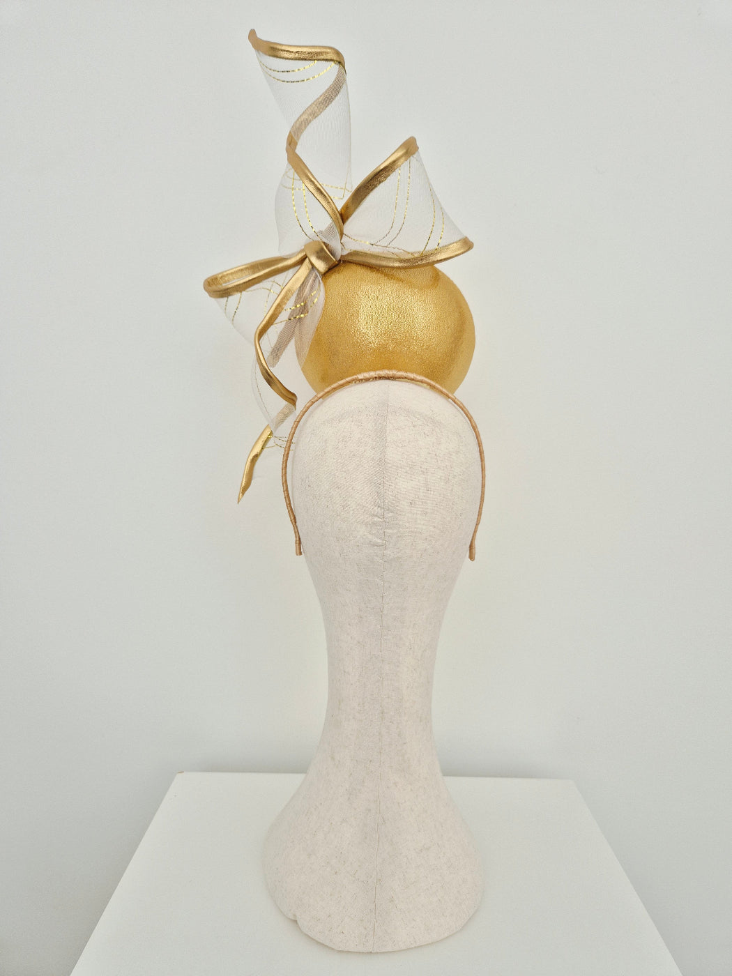 gold button percher millinery with bow and tails on a manniquin head against a white background