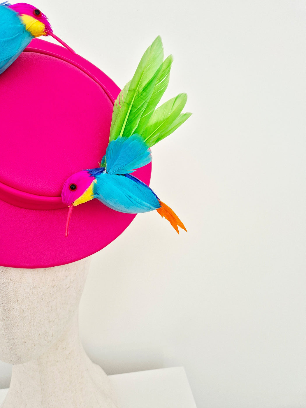 A bright pink fascinator hat displayed on a mannequin head against a clean white background, decorated with colorful bird figurines and green and blue feathers. close up