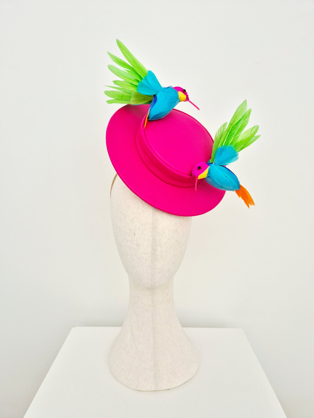 A bright pink fascinator hat displayed on a textured off-white mannequin head. The hat features two decorative bird figures with turquoise blue bodies, green wings, and orange beaks, set against a clean white studio background.
