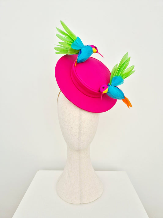 A bright pink fascinator hat displayed on a textured off-white mannequin head. The hat features two decorative bird figures with turquoise blue bodies, green wings, and orange beaks, set against a clean white studio background.