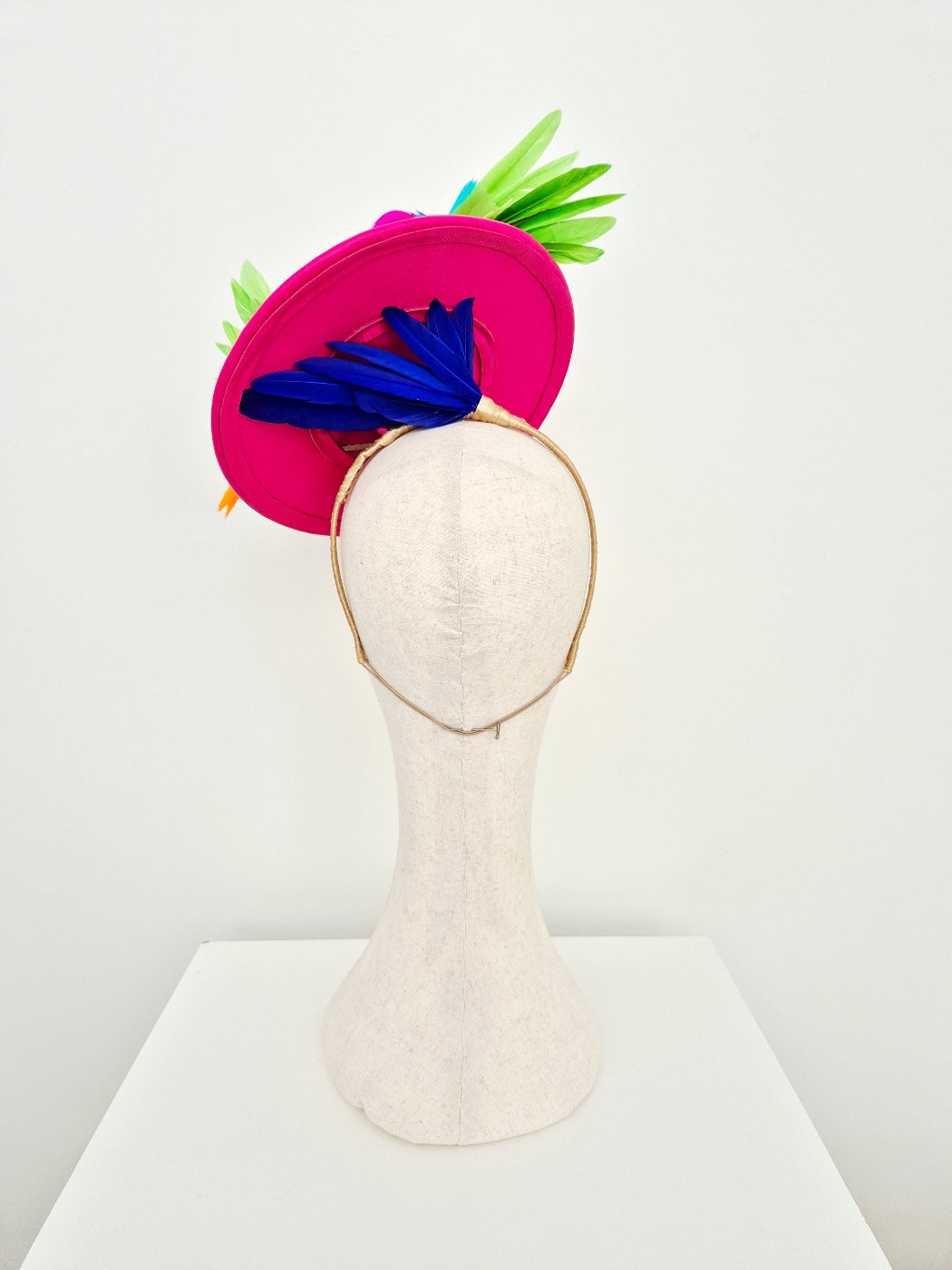 A bright pink fascinator hat with green and blue feather accents, displayed on a cream fabric mannequin head against a white background.back view