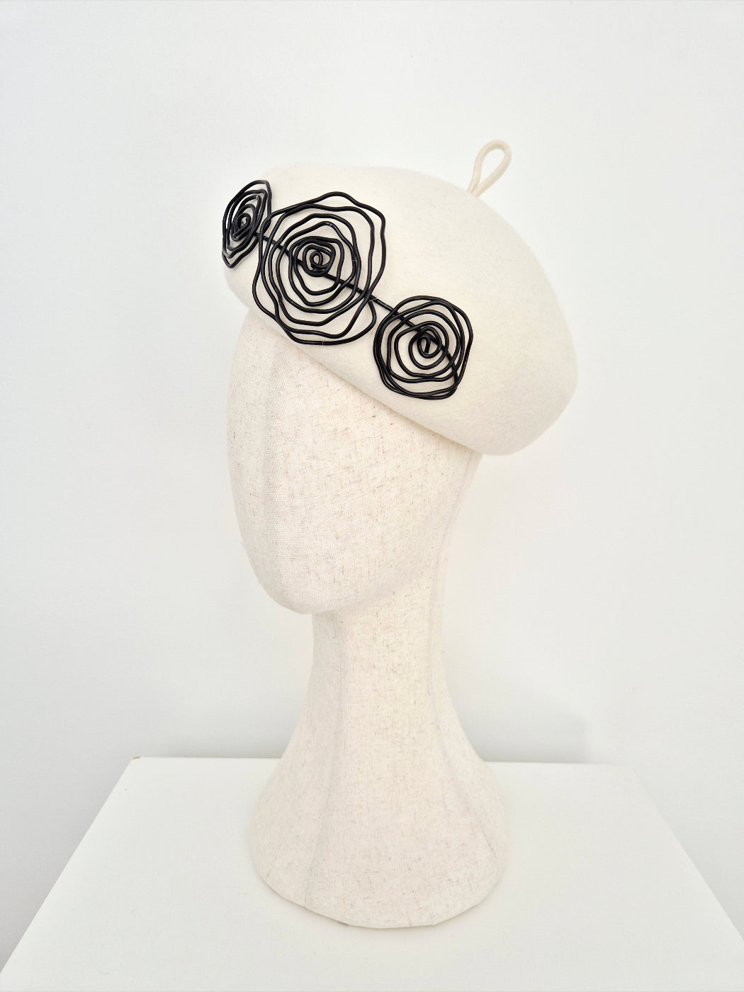 An ivory felt beret hat with three black wire rose embellishments on the side, on a  mannequin head against a white background. side view