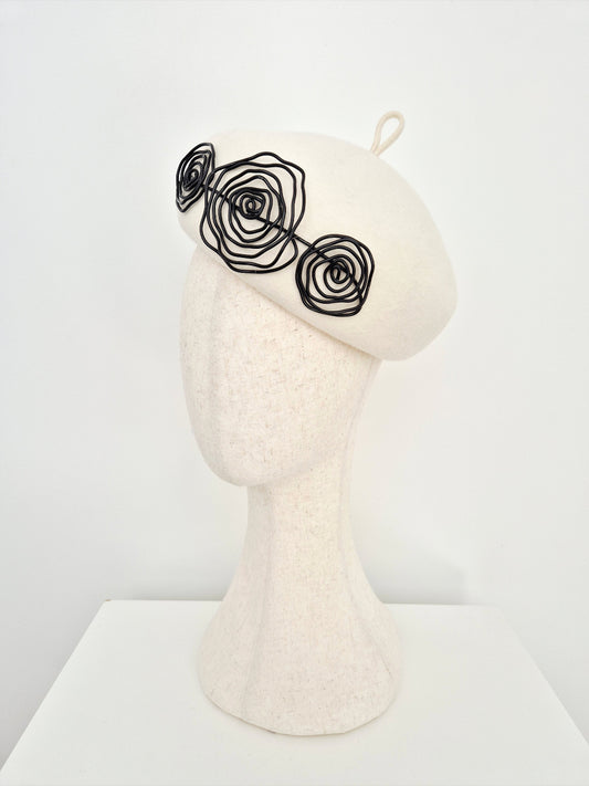 An ivory felt beret hat with three black wire rose embellishments on the side, on a  mannequin head against a white background. side view