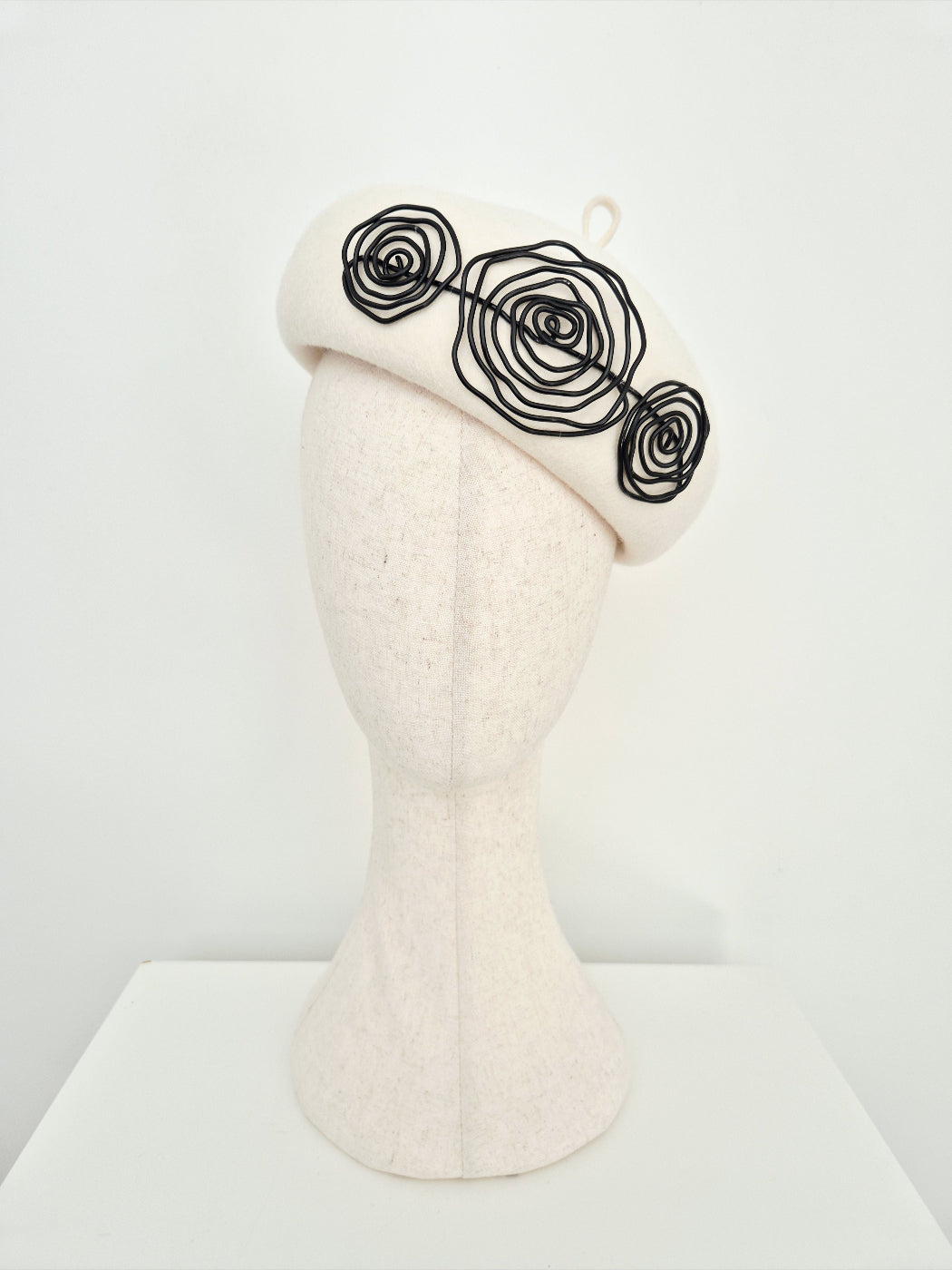 A Ivory colored felt beret with three black wire roses on a mannequin head against a white background.