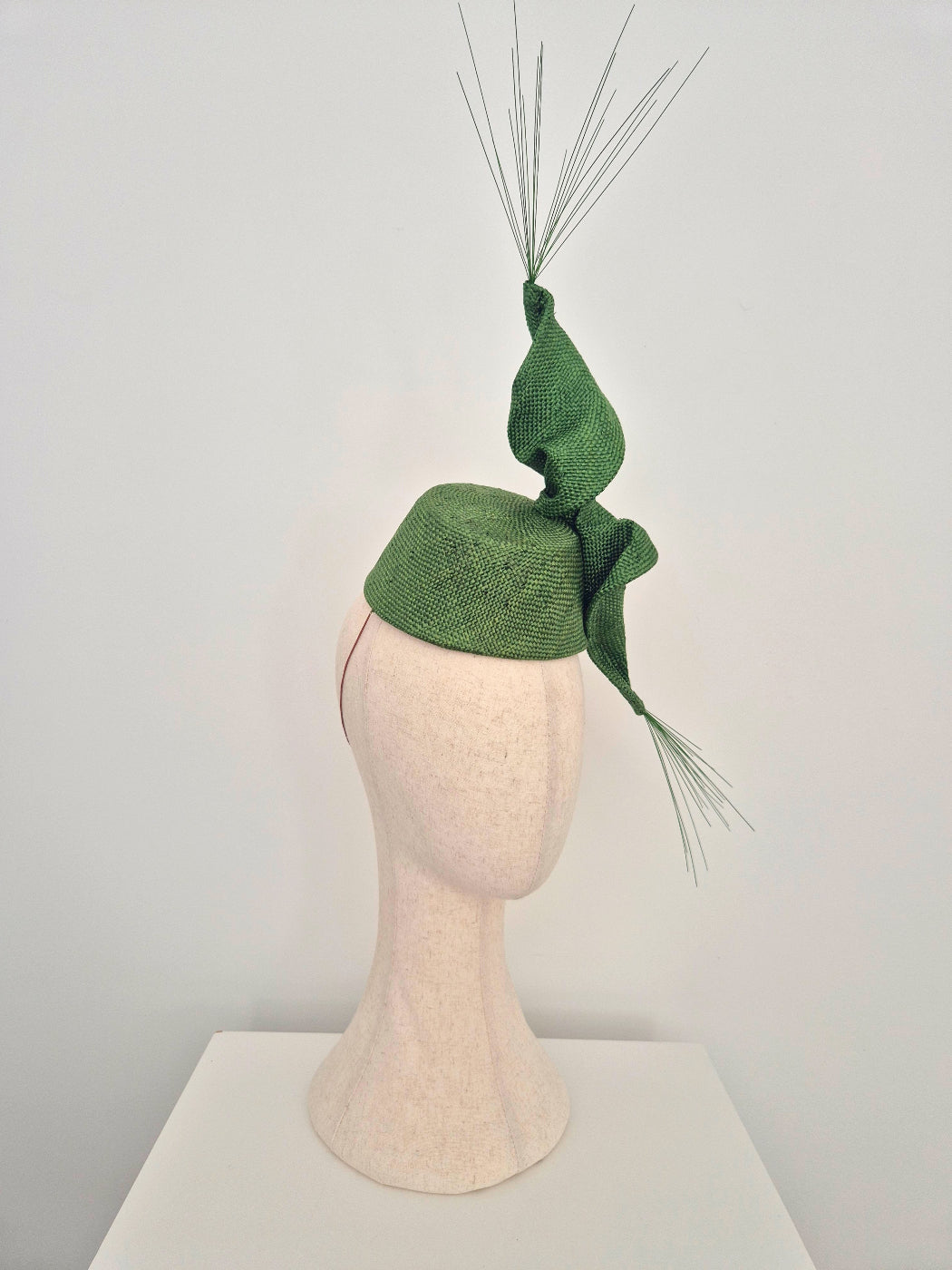 A green straw textured fascinator hat  with scrunch twist and spikes displayed on a cream-colored mannequin head against a plain light gray background.side view