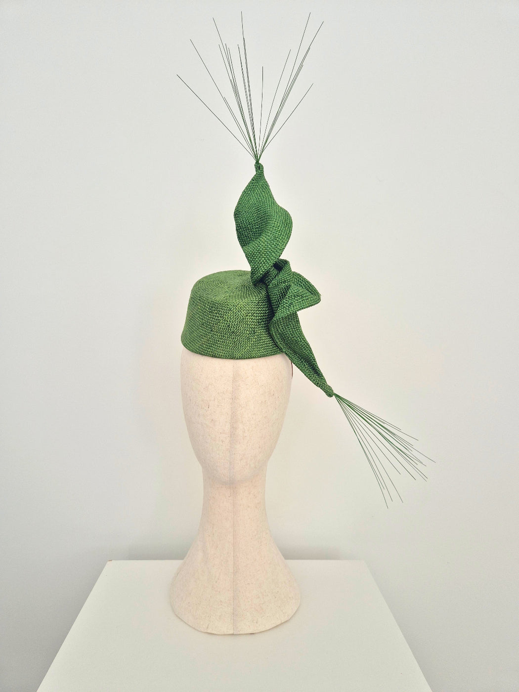 A green textured fascinator hat with a large twist and long thin spike accents, displayed on a cream-colored mannequin head against a plain light background.