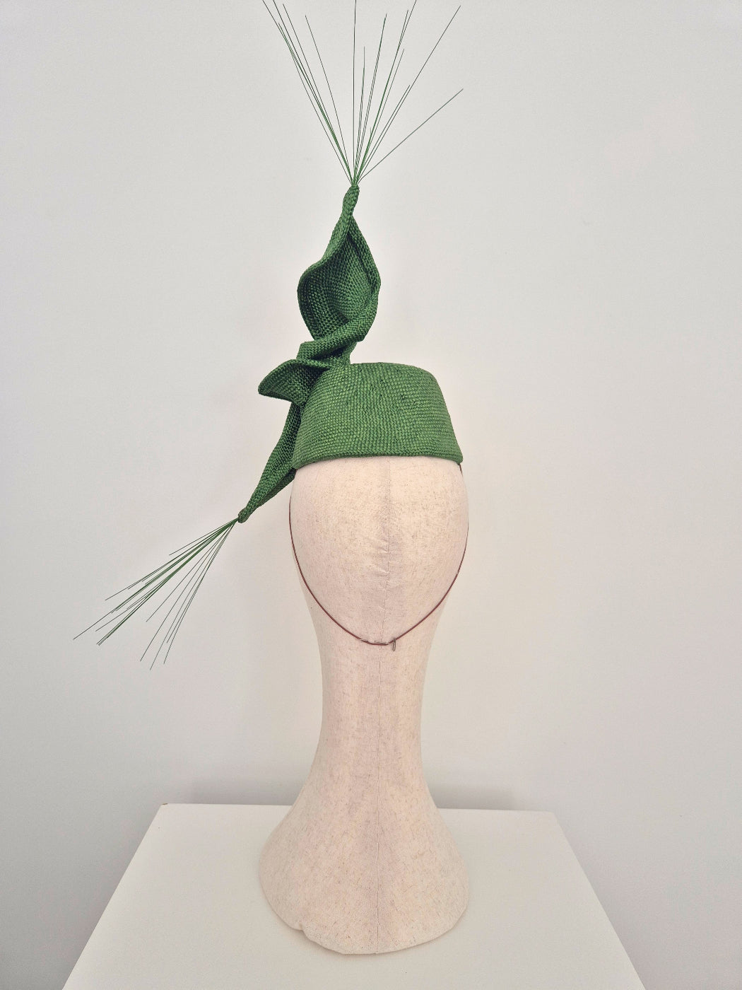 A green fascinator hat with a textured woven fabric base, a large twist, and long thin spikes displayed on a beige fabric mannequin head against a plain light gray background.back view