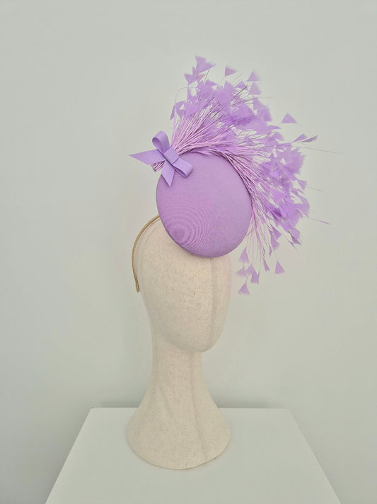 A lavender purple fascinator hat with a matching ribbon bow and delicate feather plumes, displayed on a cream-colored mannequin head against a light gray background.side view