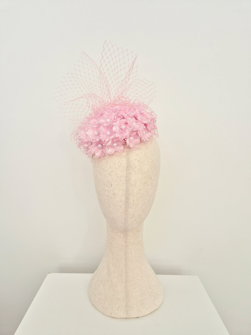 A light pink fascinator hat with a floral crown and delicate netting veil, displayed on a cream-colored felt mannequin head against a plain white background.