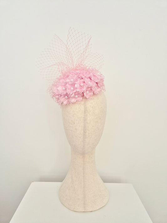 A light pink fascinator hat with a floral crown and delicate netting veil, displayed on a cream-colored felt mannequin head against a plain white background.