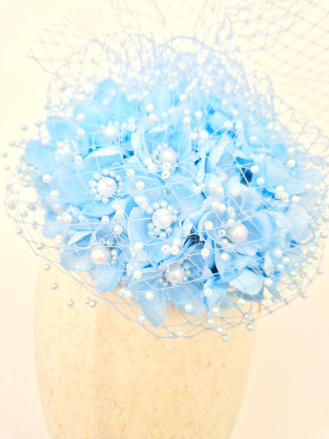 A light blue fascinator with flowers, white faux pearls, and a sheer net veil, displayed on a textured cream-colored mannequin head against a warm cream background. close up