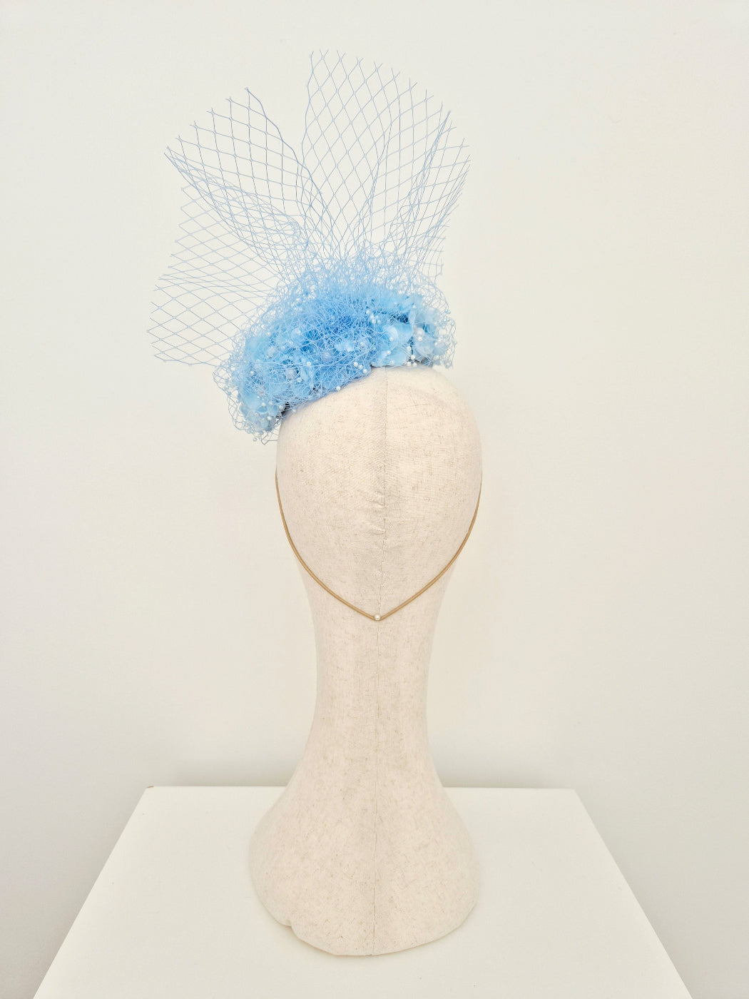 Light blue flower encrusted button headpiece with veiling back view on a manniquin head against a white wall