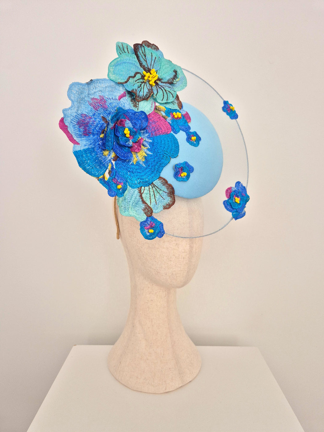 A decorative fascinator hat displayed on a beige mannequin head against a plain light background. The hat features a light blue oval base with large embroidered blue and turquoise flowers, smaller pink and blue blossoms, and a thin clear circular wire frame extending from the sides. side view