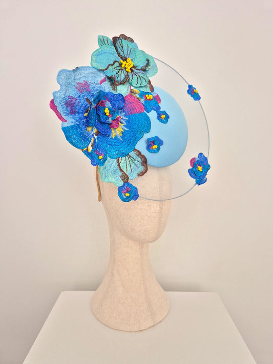 A decorative fascinator hat displayed on a beige mannequin head against a plain light background. The hat features a light blue oval base with large embroidered blue and turquoise flowers, smaller pink and blue blossoms, and a thin clear circular wire frame extending from the sides. side view