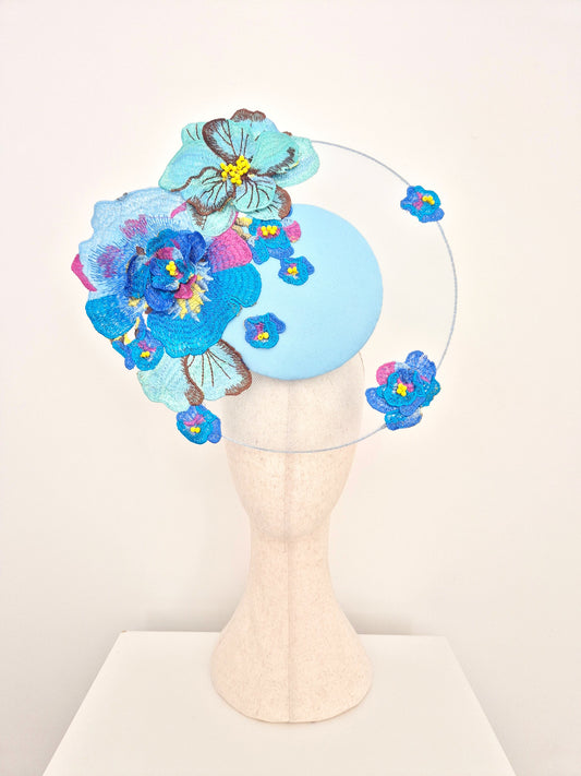 A light blue felt fascinator hat with embroidered blue, pink, and turquoise flowers, displayed on a cream-colored mannequin head against a plain white background.