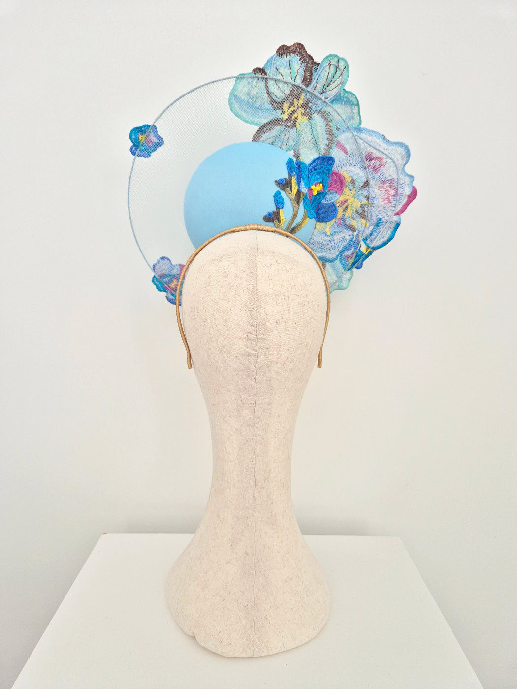 A decorative fascinator hat displayed on a cream fabric mannequin head. The hat features a light blue circular base with large embroidered blue and purple flowers, a clear wire arch, and small blue floral accents.back view